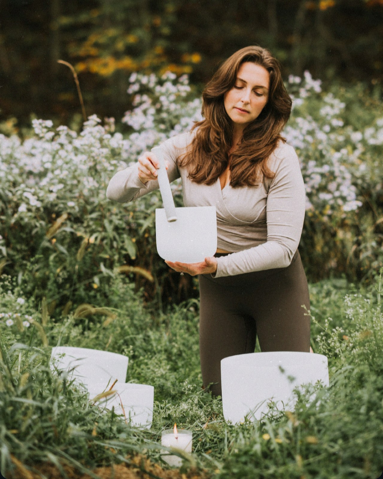 Ashley Andrews playing a crystal singing bowl in a field of white wildflowers, with two more bowls and a lit candle in the grass